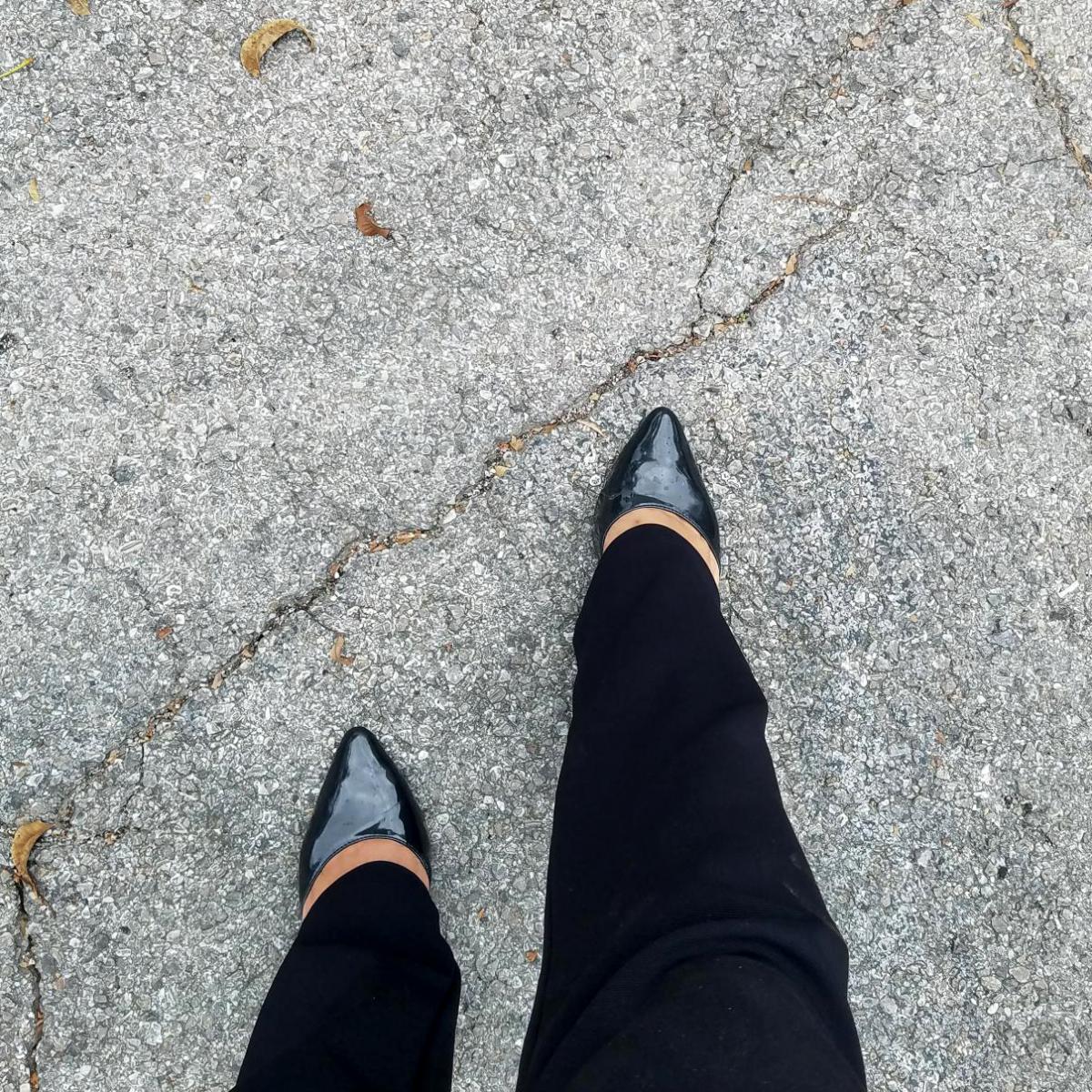 Person's feet walking on concrete wearing black pointy shoes.